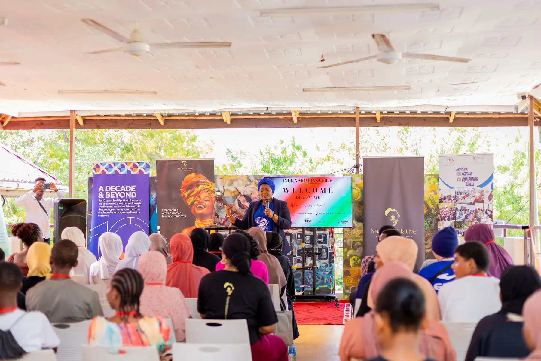 A speaker addressing seated participants in the open-sided hall